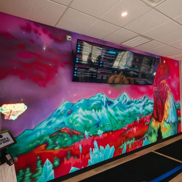 A vibrant mural of a rooster and mountains with menu screens and an "Order Here" sign at Chicks Chicken.