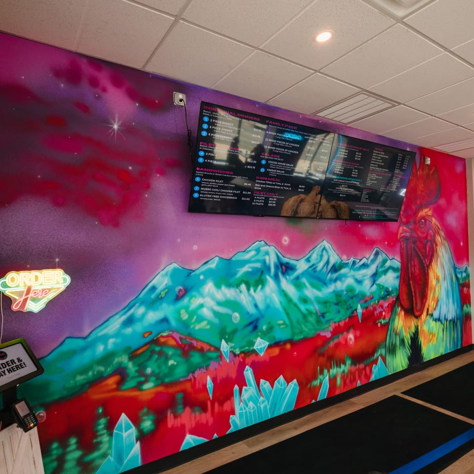 A vibrant mural of a rooster and mountains with menu screens and an "Order Here" sign at Chicks Chicken.