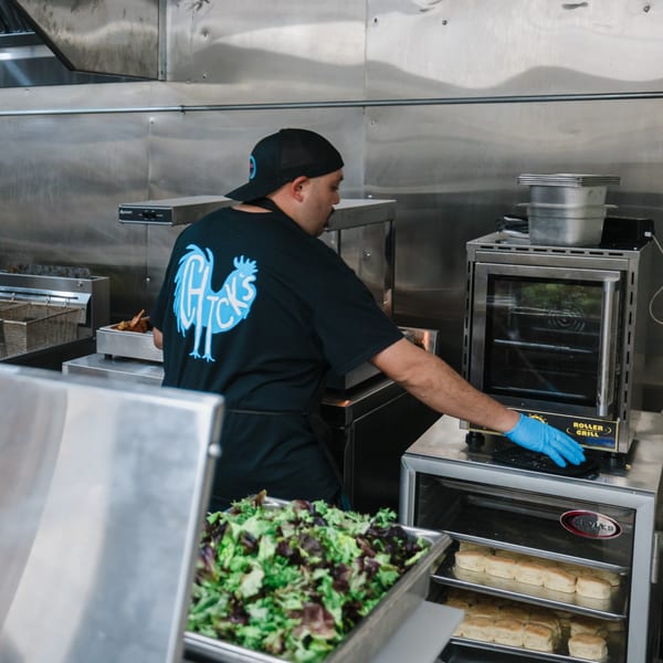 A Chicks Chicken cook in a black shirt and cap prepares food in a stainless steel kitchen.